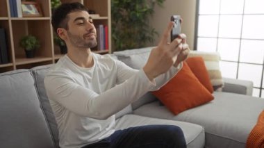 Genç adam evinde selfie çekiyor parlak oturma odasındaki koltukta turuncu yastıklar ve modern dekorlarla oturuyor..
