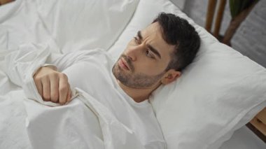 Genç adam korkmuş bir şekilde yatakta uzanıyor, kendini yatak odasının iç kısmında beyaz bir çarşafla örtüyor, evde duygusal, kapalı bir ortam izlenimi uyandırıyor..