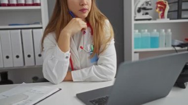 Üniformalı genç beyaz kadın doktor. Parmakları çenesinde düşünüyor. Klinikte şaşkınlık ve şüphe içinde başını kaldırıyor.