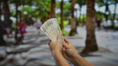 Bulanık insanlar ve palmiye ağaçları ile çevrili bir şehir caddesinde elinde para tutan adam, hareketli bir şehir ortamında finansal bir alışverişi vurguluyor..