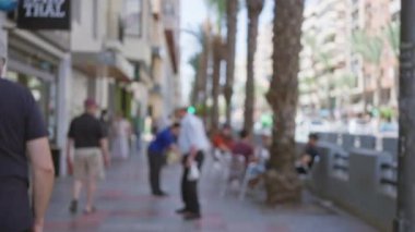 Güneşli bir günde açık bir şehir ortamında palmiye ağaçları ve binaların yanından geçen kadın ve erkeklerin olduğu bulanık sokak sahnesi.