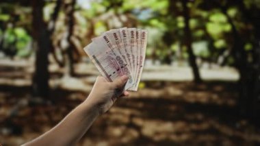 Suudi riyal banknotlarını elinde tutan adam parkın dışında, doğal bir bahçe arka planına karşı para sergiliyor..
