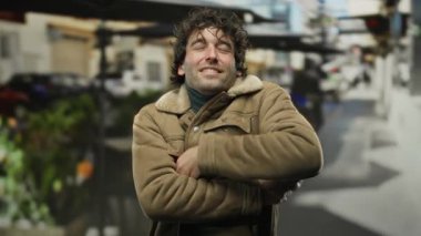 İspanyol bir adam, şehir sokaklarında kahverengi bir ceket giyerek, soğuk bir şekilde, gözleri kapalı, çekici ve yakışıklı bir görünüm sergileyerek kendini kucaklıyor..