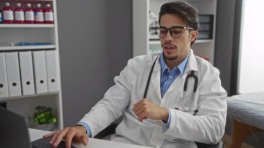 Klinik ortamında steteskop takan genç bir adam, iş yerinde bilgisayarla meşgul, tıbbi malzemeler ve organize dosyalarla çevrili..