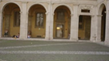 Üniversite kampüsünün avlusunda insanların, kemerlerin ve tarihi mimarinin bulanık görüntüsü, açık hava ortamında huzurlu bir akademik atmosfer taşıması.