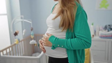 Yatak odasında beşiği olan hamile bir kadın, karnını nazikçe tutuyor, huzurlu ve rahat bir ortamda yakın bir anneliği ima ediyor.