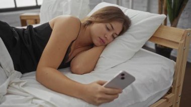 Woman lying on a bed in a bedroom, holding a smartphone, showcasing a serene interior home setting and reflecting a relaxed lifestyle