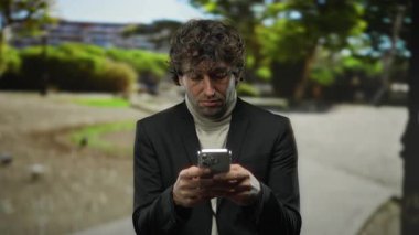 Şehir caddesinde akıllı telefon kullanan, siyah takım elbiseli ve balıkçı yakalı bir adam, bulanık şehir yeşiliyle çevrili, açık havada ciddi bir yüz ifadesi sergiliyor..