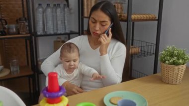 Kadın, oyuncaklarla ve mutfak eşyalarıyla çevrili rahat bir apartman dairesinde bebeğini tutarken telefonda birden fazla iş yapıyor..