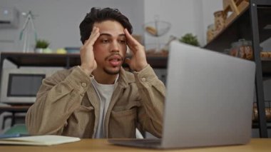 Ofiste çalışan genç adam, iş yerindeki gerginliği yansıtan yakınlarda dizüstü bilgisayarla başını tutarak stresli bir şekilde çalışıyor.