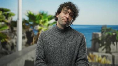 Kazak giymiş kıvırcık saçlı genç adam güneşli bir günde sahilde dikiliyor. Arka planda palmiye ağaçları ve plajda rahat bir hava var..