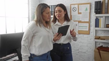 Ofiste çalışan kadınlar dijital tablet kullanıyor, proje fikirlerini tartışıyor, gündelik kot pantolon giyiyor, modern çalışma istasyonu ve ilan panosundaki belgelerle çevrili..