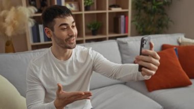 Parlak bir oturma odasındaki İspanyol genç adam akıllı bir telefonla konuşuyor. İçerideki rahat bir kanepede heyecanını ifade ediyor ve gülümsüyor. Ev dekorasyonu ve kitaplıklarla dolu..