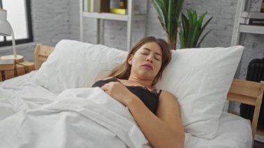 Beyaz çarşaflar ve yumuşak yastıklarla sıcak bir yatak odasında isteksizce uyanan bir kadın yorgun bir yüz ifadesi sergiliyor. Sabahın erken saatlerinde modern ve şık bir iç mekanda rahatsız olduğunu gösteriyor..