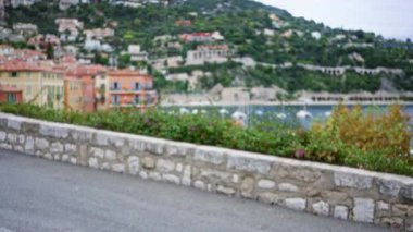 Villefranche-sur-mer 'daki açık hava sahnesinde bulanık arka planda renkli evler, yeşil tepeler ve taş bir duvar, sakin ve resimli bir Fransız sahil kasabasını yakalıyor..