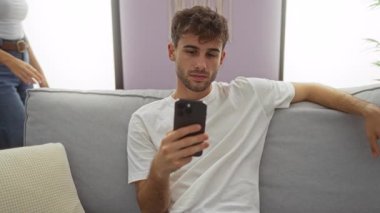 Kanepede oturan erkek ve kadın rahat oturma odasında akıllı telefon kullanıyor, kapalı alanda sevgi dolu bir anı paylaşıyor, rahat ev atmosferini modern dekor ve günlük kıyafetlerle tasvir ediyorlar..