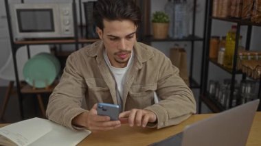 Gündelik kıyafetli genç adam modern ofis ortamında telefonunu kontrol ederken dizüstü bilgisayarda çalışıyor..