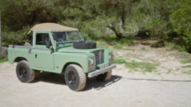 Ormanlık bir kampta park etmiş yeşil askeri araç parlak güneş ışığı altında arka planda dağılmış ağaçları ve engebeli arazisiyle kırsal ve maceraperest bir his veriyor..