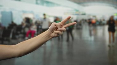 Yoğun bir havaalanında farklı el hareketleri sergileyen bir adam, bulanık bir kalabalığın önünde rakamları gösterir, seyahat beklentisi ve iletişimi yansıtır..