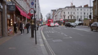Sonbaharda kalabalık Londra caddesi, ikonik kırmızı otobüsler de dahil olmak üzere yürüyen insanlarla dolu. Bulanık odaklı, atmosferik derinliği artıran tipik bir şehir manzarası yaratıyorlar..