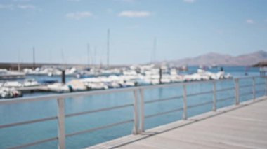 Bulanık marina arka planı, açık mavi gökyüzü ve uzak dağlarla limana yanaşmış yatları ve tekneleri gösteriyor.