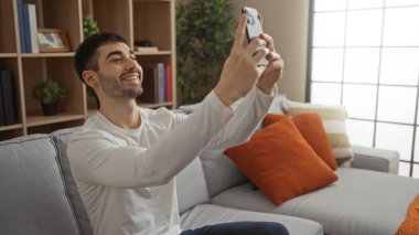 Oturma odasında selfie çeken genç bir adam. Parlak dekorlu, gülümseyen, rahat yüzlü, evde boş zaman geçirdiğine işaret eden rahat bir ortam sergiliyor..