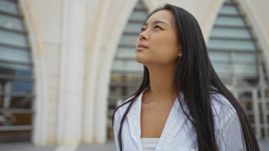 Genç bir kadın şehir merkezinde dikiliyor ve arka planda modern bir şehir binası var.