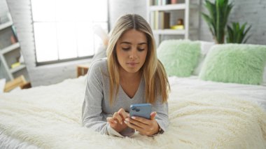 Rahat yatak odasında rahat bir kadın akıllı telefon kullanıyor, yumuşak yatağa uzanıyor, rahat ve sakin bir havası olan sarışın, aydınlık ve davetkar bir şekilde evde boş vakitlerin tadını çıkarıyor..