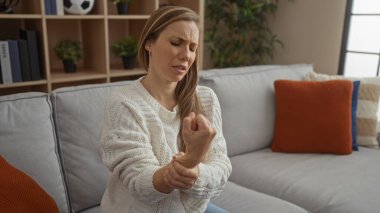 Oturma odasındaki gri bir kanepede oturan bir kadın bileğini tutarak kederli bir yüz ifadesiyle rahat bir ev ortamında yastıklar ve bitkilerle çevrili..