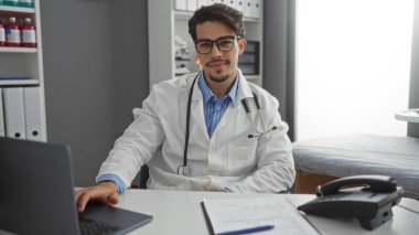 Klinikteki genç adam laboratuvar önlüğü giyiyor, dizüstü bilgisayarda çalışıyor, profesyonel ve kendinden emin görünüyor, steteskop ve dosya ile hastane masasında..