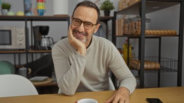 Ofis ortamında güvenle gülümseyen orta yaşlı bir adam, olgun ve yakışıklı bir görünüm sergiliyor, düşünceli bir ifadeyle, iş yerinde evde oturuyor..