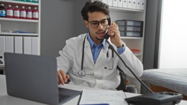 Genç İspanyol doktor, klinikte telefonla konuşuyor. Bilgisayarlı steteskop kağıtları rafları ve çalışma alanı profesyonel bir ortamda görülüyor.