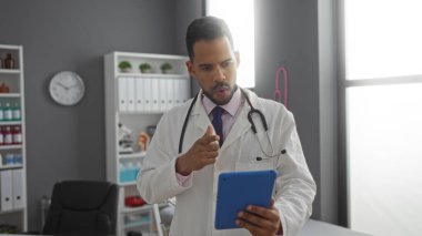 Klinikte elinde tablet olan ve steteskop takan bir adam ekranda dikkatlice görünüyor. Bu da hastane ortamında profesyonel bir odak noktası olduğunu gösteriyor..