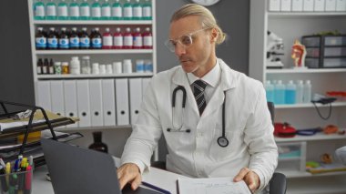 Klinik odasında orta yaşlı beyaz bir adam steteskop takıyor, masasında tıbbi malzeme ve raflarla çevrili bir ifadeyle belgeleri inceliyor..