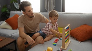 Baba ve küçük oğlu koltukta renkli bir oyuncakla oynuyorlar rahat bir evde oturma odası düzeninde, yastıklar ve doğal ışıklarla çevrili bir aile ortamında..