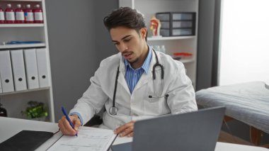 Genç İspanyol doktor, klinik odasına dizüstü bilgisayar, steteskop ve tıbbi dosyalarla notlar yazıyor..