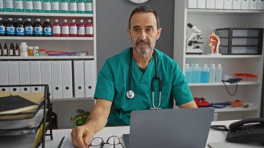 Yeşil önlüklü İspanyol erkek doktor, tıbbi ekipmanlarla organize bir klinikte oturuyor ve profesyonel ve sakin bir sağlık hizmeti ortamı sergiliyor..