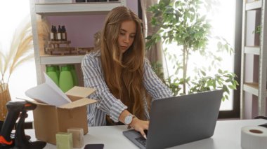 Dekoratif eşyalar ve bitkilerle çevrili bir ev dekorasyonu mağazasında dizüstü bilgisayarda çalışan genç bir kadın bir kutuyu işaretliyor ve çevrimiçi teslimat için bir sipariş hazırlıyor..