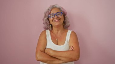 Gri saçlı ve gözlüklü İspanyol bir kadın kollarını kavuşturarak güvenle gülümsüyor, izole edilmiş pembe bir arka plana karşı duruyor, orta yaşta güzellik ve zarafet sergiliyor..