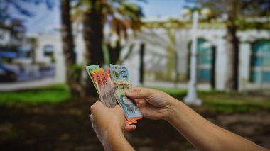 Arka planında yeşil park olan güneşli bir şehir sokağında Avustralya doları tutan adam döviz kurunu veya açık havada para transferini sergiliyor..