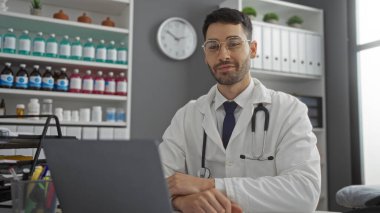 Laboratuvar önlüğü ve gözlükleriyle klinikte oturan ve arka plandaki raflarda duran İspanyol asıllı genç adam ve kapalı alandaki kameraya bakan steteskop.