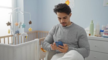 Genç bir adam, beşiğin yanındaki bir yatak odasında akıllı telefon kullanarak gün içinde sıcak ve davetkar bir şekilde rahat bir atmosfer yaratıyor..