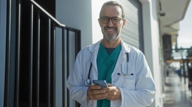 Tıbbi ceketli İspanyol bir adam, şehir sokağında gülümsüyor. Akıllı telefon kullanıyor. Hastanenin yanında kendinden emin görünüyor..