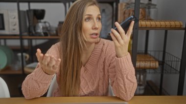 Ofis ortamında akıllı telefondan konuşan bir kadın. Etrafı raf ve mutfak aletleriyle çevrili. El hareketleriyle duygularını ifade ediyor..
