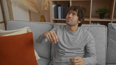 Modern apartmandaki koltukta oturan Latin kökenli genç adam rahat yastıklar ve oturma odasında onu çevreleyen dekorasyonla derin derin düşünüyor..