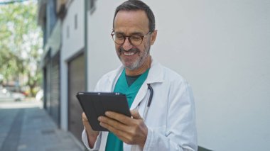 Beyaz önlüklü ve gözlüklü İspanyol doktor, hastanenin yanındaki güneşli bir şehir sokağında dijital tablet kullanırken gülümsüyor..