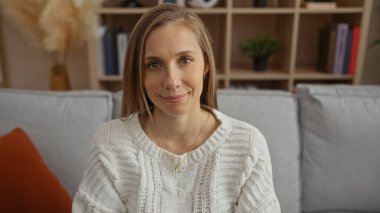 İçerde oturan kadın, rahat bir oturma odasında gülümsüyor, beyaz bir kazak giyiyor, ev dekorasyonu elemanlarıyla çevrili, sıcaklık ve rahatlık taşıyor..