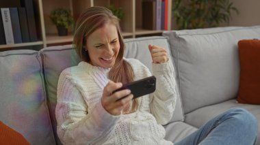 Kadın evde yumruğunu sıkarak kutlama yapıyor. Oturma odasındaki koltukta telefona bakıp gülümsüyor. Evde neşe ve heyecan ifade ediyor..