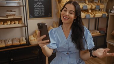 İspanyol kadın, arka planda ekmek rafları olan bir pastanede görüntülü konuşma yapıyor.