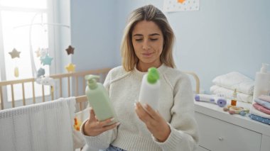 Konforlu bir yatak odasında şişeleri inceleyen kadın, arkasında beşiği olan bebek aksesuarlarıyla çevrili iki yeşil ve beyaz şişe tutuyor ve pencereden güneş ışığı süzülüyor..